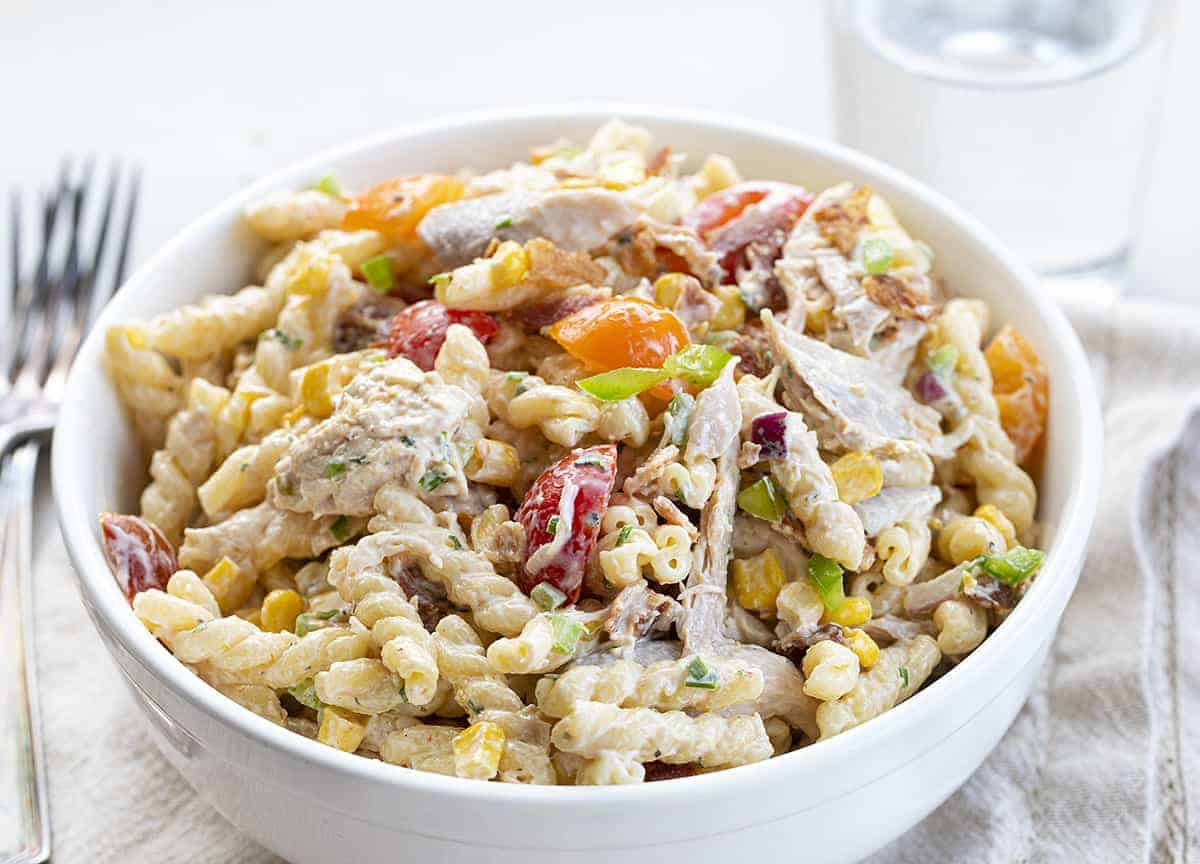 Chicken Bacon Ranch Pasta Salad is a creamy and refreshing cold pasta salad that is loaded with shredded chicken, vegetables, and crispy bacon seasoned with homemade ranch seasoning. Pasta Salad, Chicken Salad, Deluxe Chicken Salad. Ranch Chicken Salad, Chicken Bacon Ranch Salad, Bacon Chicken Ranch Salad, Cold Pasta Salad, BBQ, potluck, summer salads, recipes, side dishes, party recipes, i am homesteader, iamhomesteader BIg Bowl of Chicken Bacon Ranch Salad on White Towel with Water and Forks