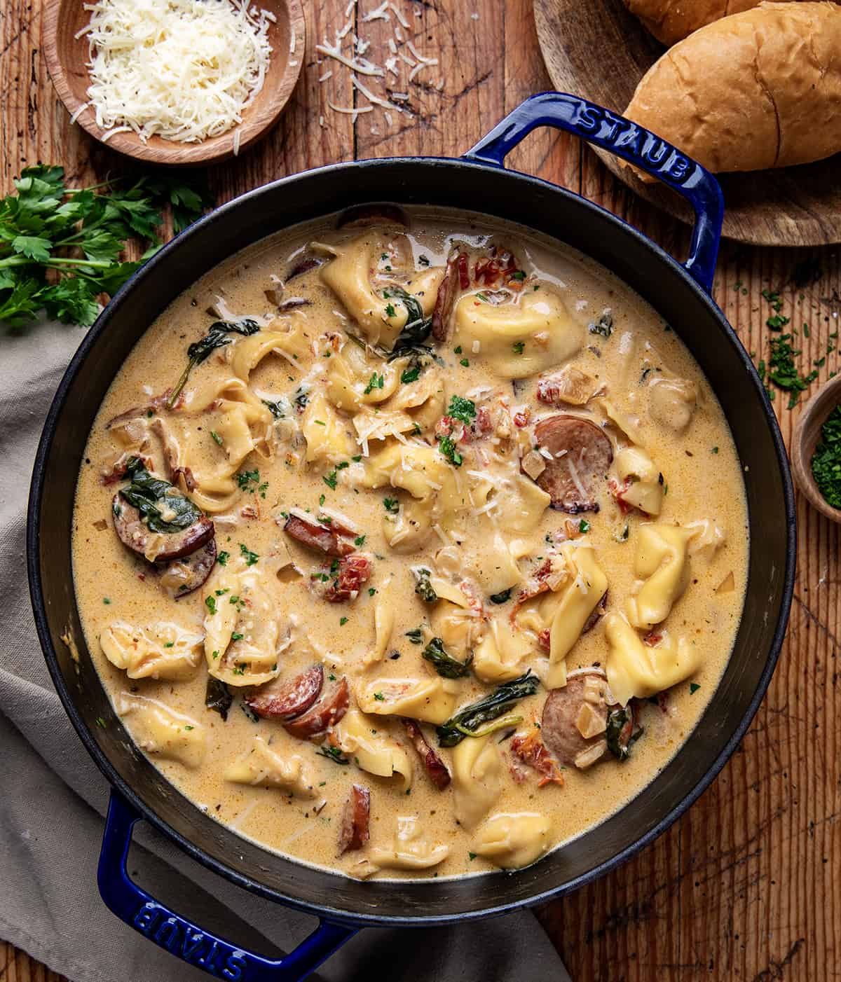 Kielbasa Tortellini Soup is a creamy broth-based soup filled with Kielbasa, sun-dried tomatoes, cheese tortellini, and fresh spinach. It's hearty enough to fill you up as well as warm you up on a cold day. Pot of Kielbasa Tortellini Soup on a wooden table from overhead.