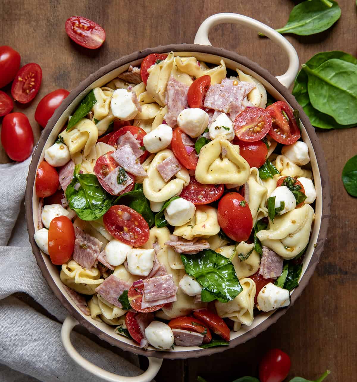 Tortellini Pasta Salad features cheese-filled tortellini with tomatoes, red onions, mozzarella, salami, and spinach, topped with a tangy olive oil and vinegar dressing. It is delicious as a lighter main dish, for lunch, or served as a side dish at potlucks, barbecues, or any casual gathering! Bowl of Tortellini Pasta Salad on a wooden table from overhead.