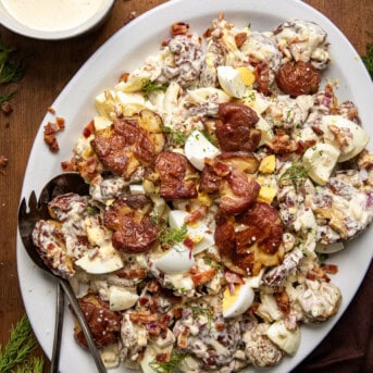 Platter of Smashed Roasted Potato Salad on a wooden table showing the smashed potatoes, hard boiled eggs, crispy bacon bits, finely diced onion, and perfect dressing that brings it all together.
