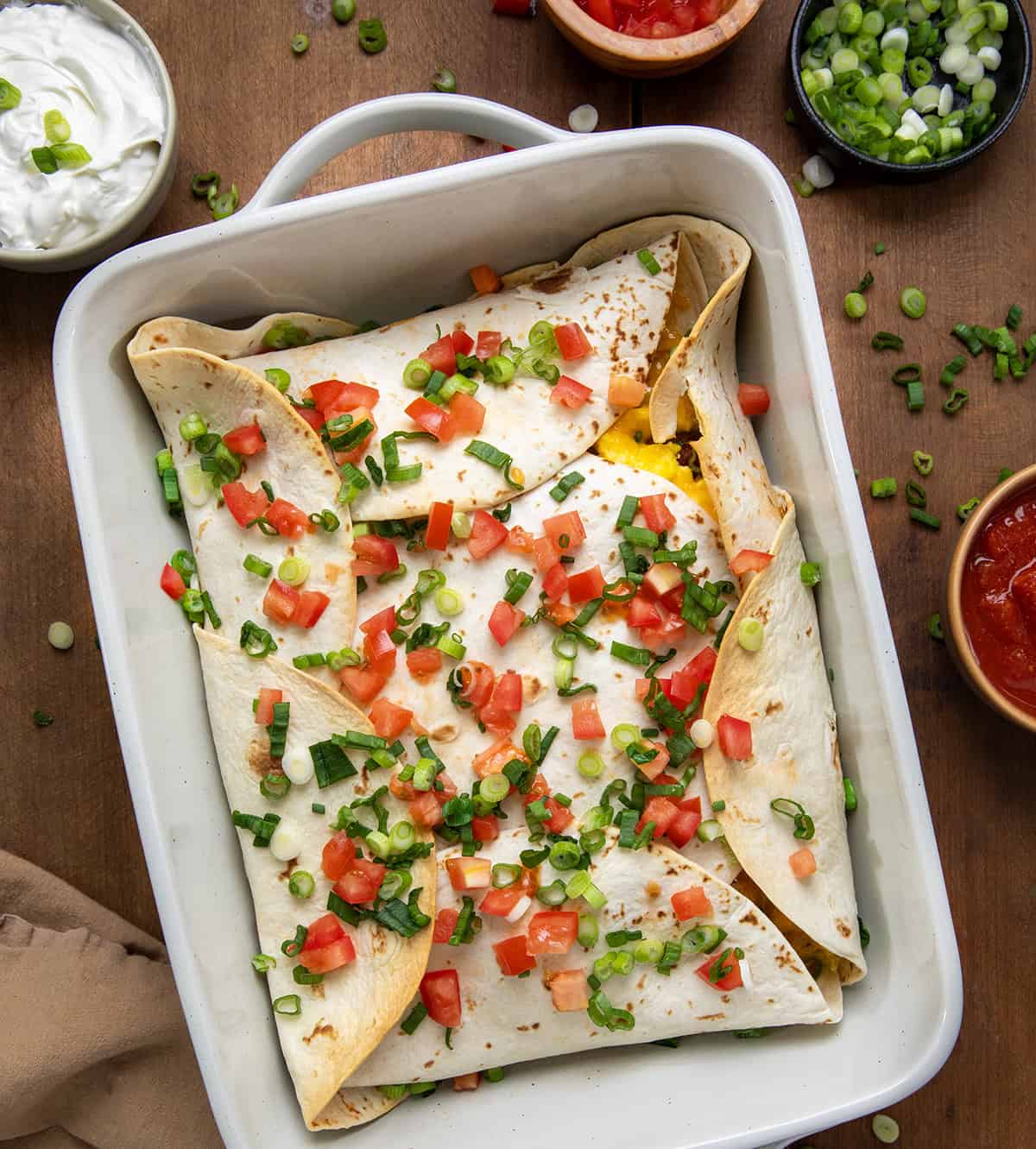 Pan of Breakfast Wrap on a wooden table surrounded by tomatoes, green onion, salsa, and napkin from overhead.