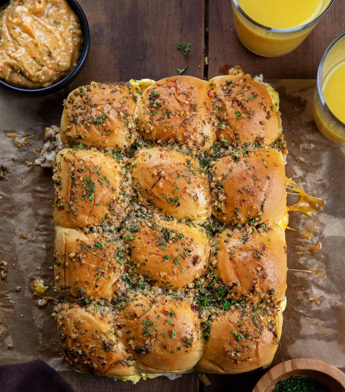 Start your day with flavor-packed Cowboy Breakfast Sliders! They are delicious mini breakfast sandwiches filled with scrambled eggs, sausage, bacon, and cheddar cheese on Hawaiian rolls! Looking down at Cowboy Breakfast Sliders on a wooden table with orange juice and cowboy butter.