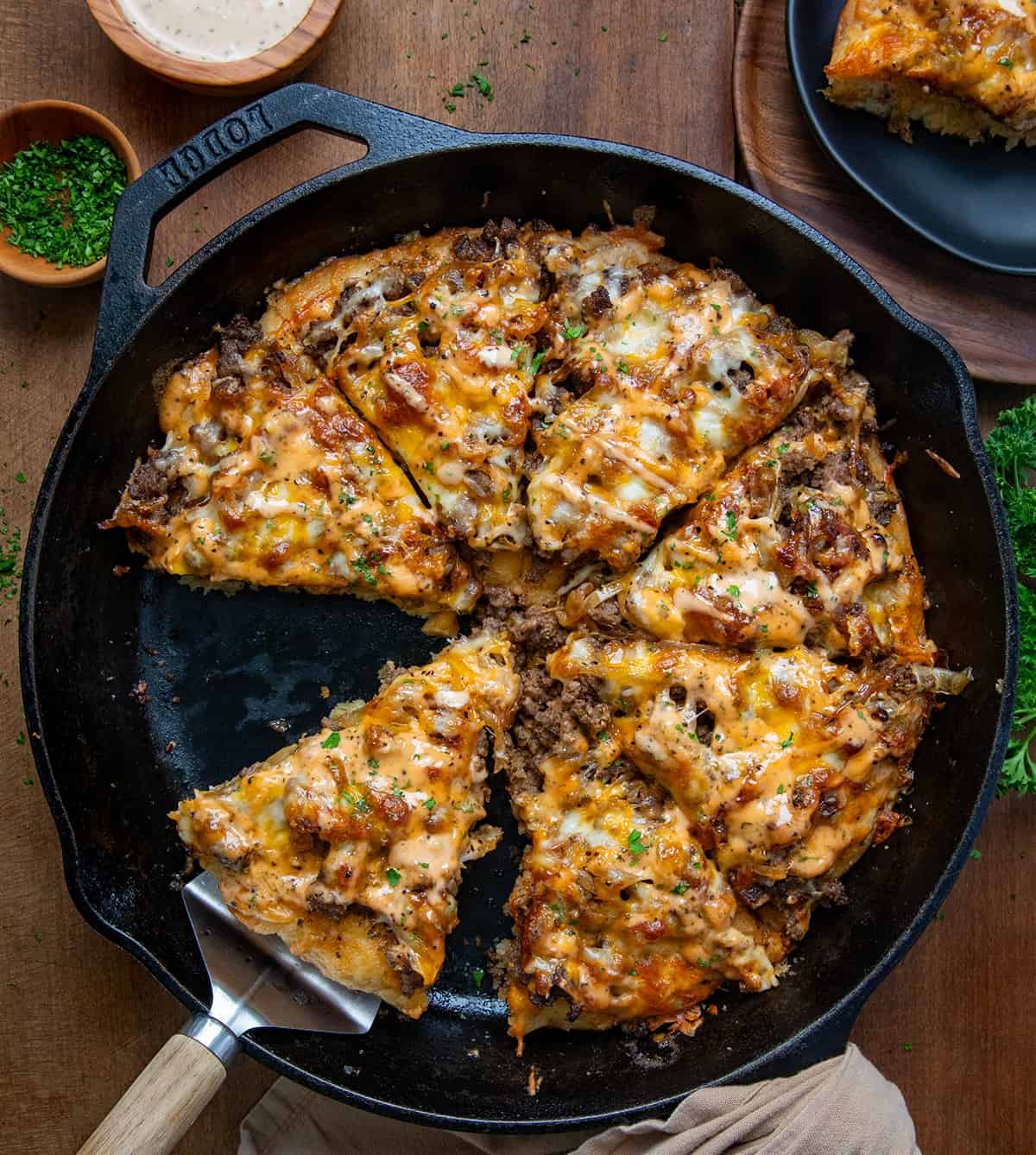 This WOW Patty Melt Pizza is my favorite focaccia bread, topped with a tangy mayo-ketchup sauce, followed by seasoned ground beef, caramelized onions, and a blend of mozzarella and cheddar cheese. Then, once baked, more sauce is drizzled over the top of the pizza or served on the side for dipping. Each bite gives you all of the delicious flavors of a patty melt in a deep dish pizza form! Patty Melt Pizza in a skillet cut into pieces and with a serving spoon in the pan.